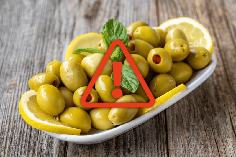 Alt text: Green olives with lemon slices on a dish, warning sign overlayed.