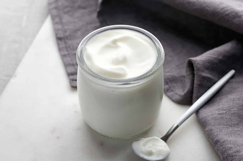 A jar of fresh Greek yogurt with a spoon on a white surface, showcasing a nutritious and delicious dairy snack.