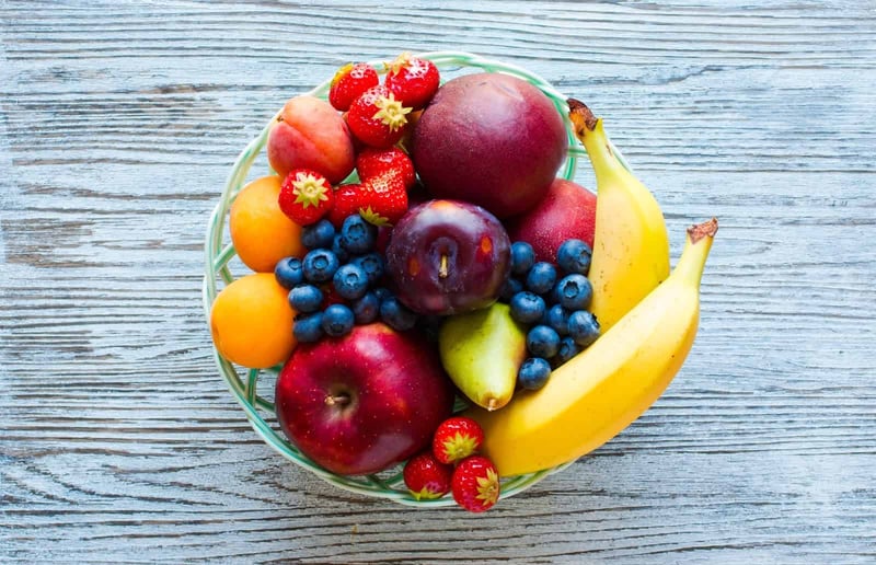 Bright assortment of strawberries, blueberries, apples, bananas, peaches, and plums in a basket on a wooden table.