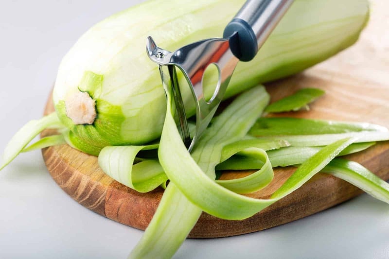 Close-up of a stainless steel vegetable peeler peeling a fresh cucumber on wooden cutting board.