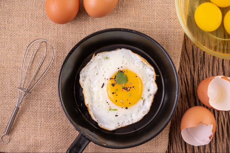 Freshly cooked sunny side up egg on a black plate, with eggshells and raw eggs nearby, ready for breakfast.
