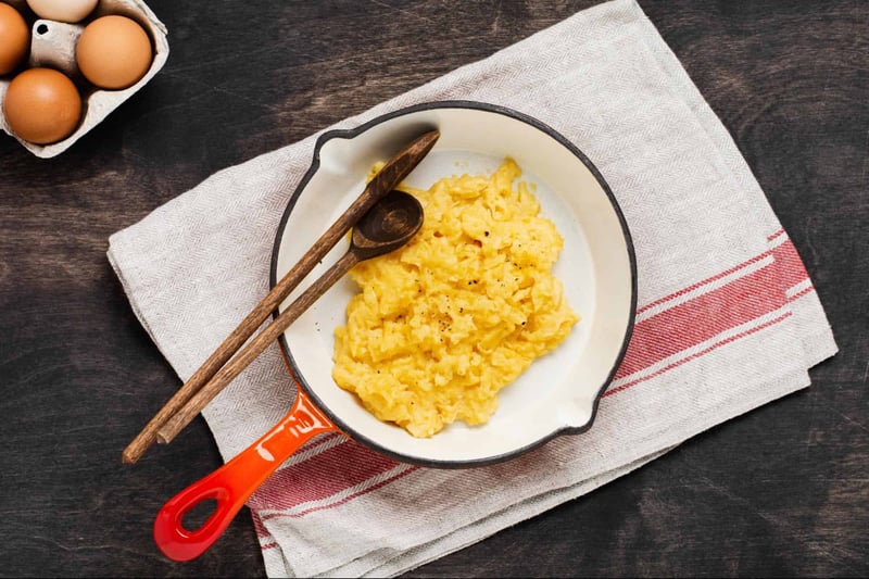 Scrambled eggs served in a skillet with wooden utensils on a white cloth napkin.