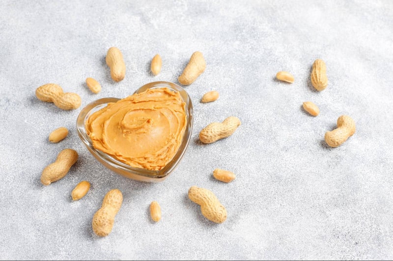 Peanut butter in a glass bowl surrounded by peanuts on a gray textured background.