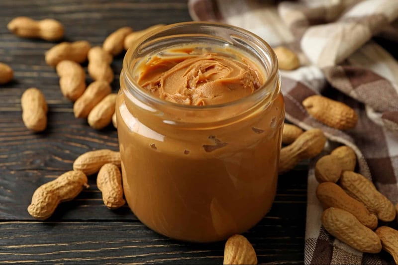 Natural peanut butter spread in a glass jar with scattered peanuts on a rustic wooden surface.