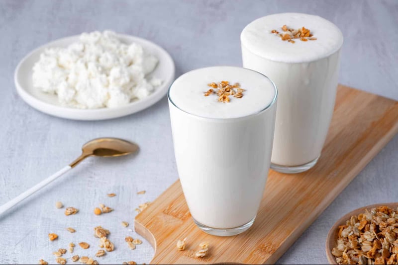 Bright image of two glasses of milk topped with oats, with oats scattered on a white surface and a plate of oats in the background.