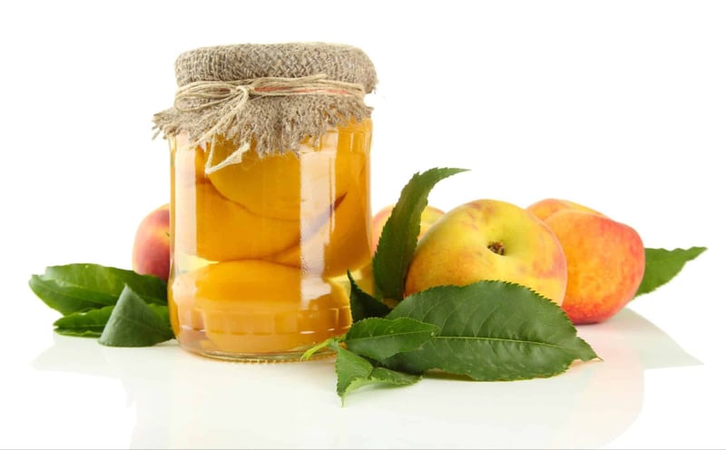 Fresh peach jam in a glass jar with a rustic fabric cover and string, surrounded by ripe peaches and green leaves.
