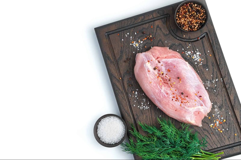 Fresh raw chicken breast seasoned with salt and pepper on a wooden cutting board with herbs and spices.