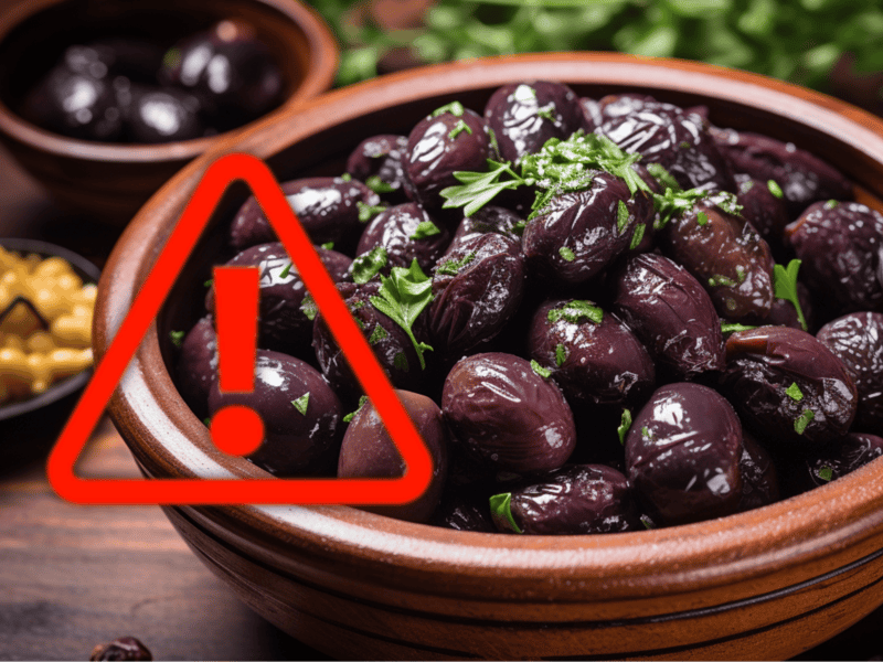 Close-up of ripe black olives in a bowl, with a red warning icon overlay.