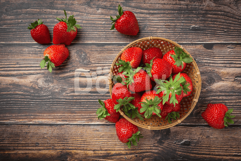 Bright red strawberries with green leaves in a wicker basket on rustic wooden table.