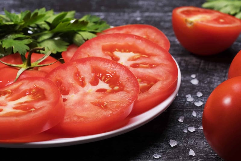 Bright, fresh tomatoes sliced for salads or cooking, on dark rustic wood with herbs and salt.