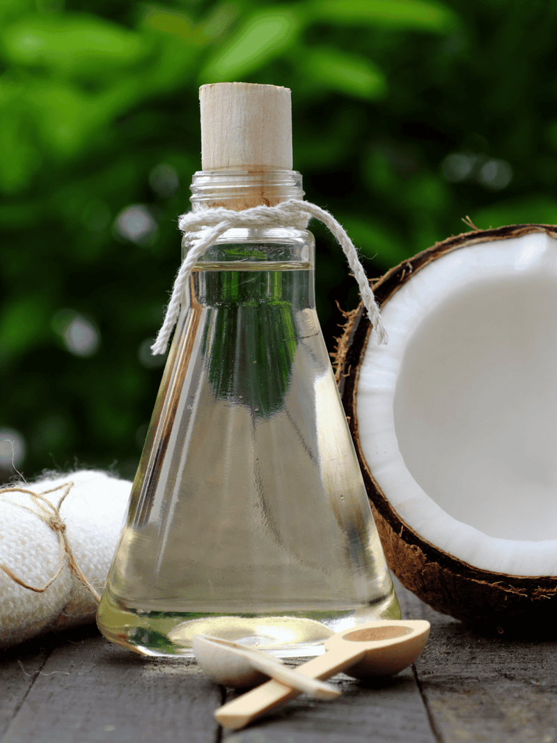 Coconut oil in a glass bottle with coconut halves and spoons on a rustic surface.