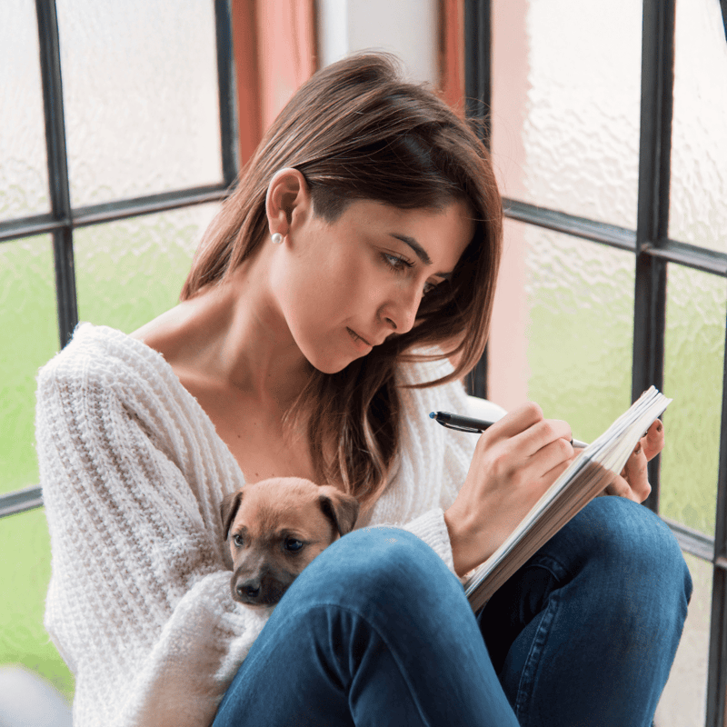 Woman with a puppy, sitting by the window, journaling or working on a project peacefully.