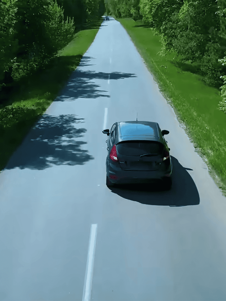 Aerial view of a black car traveling down a scenic rural road with lush green trees on both sides.