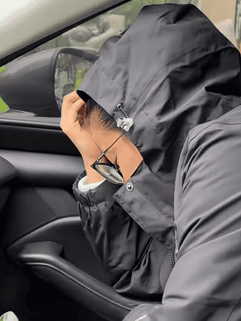 Person in black raincoat covering face with hand in vehicle.