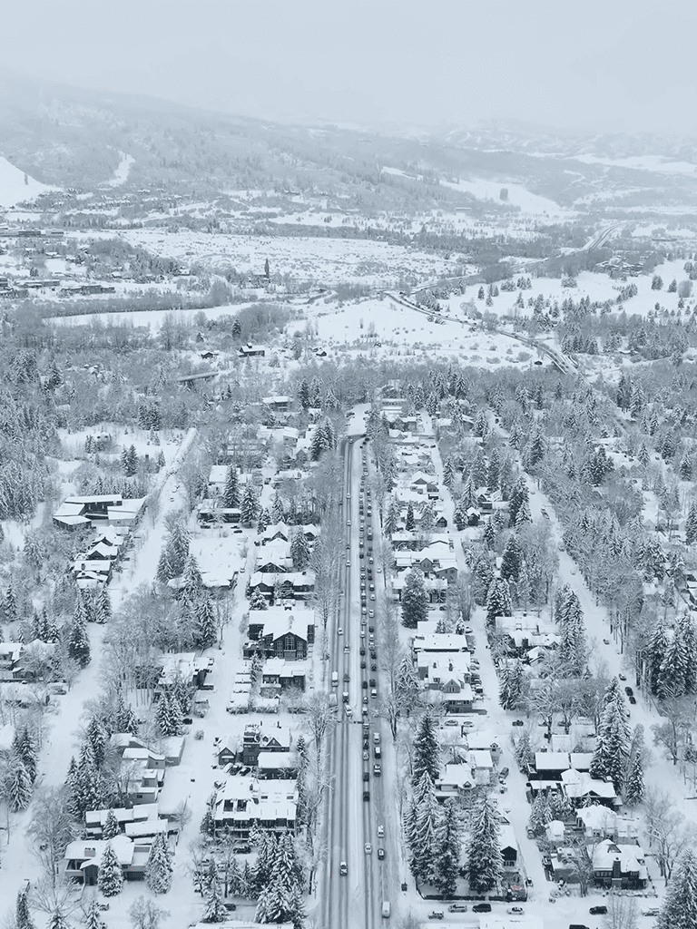 Snowy suburban neighborhood with snow-covered houses, trees, and a busy traffic-filled road in winter.