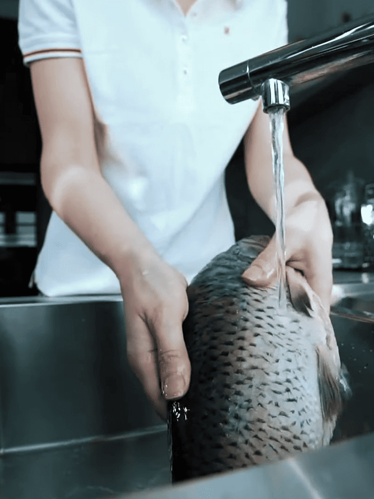 ALT: Person cleaning fish under running water in a kitchen sink.