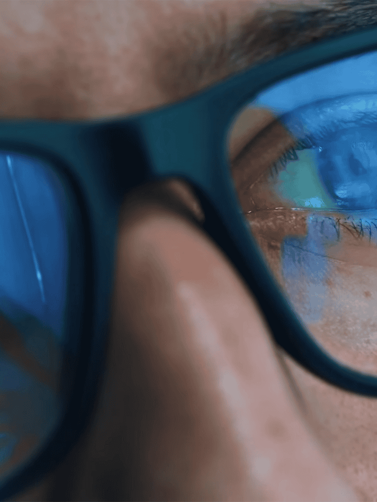 Close-up of a person wearing glasses reflecting a screen with blue light.