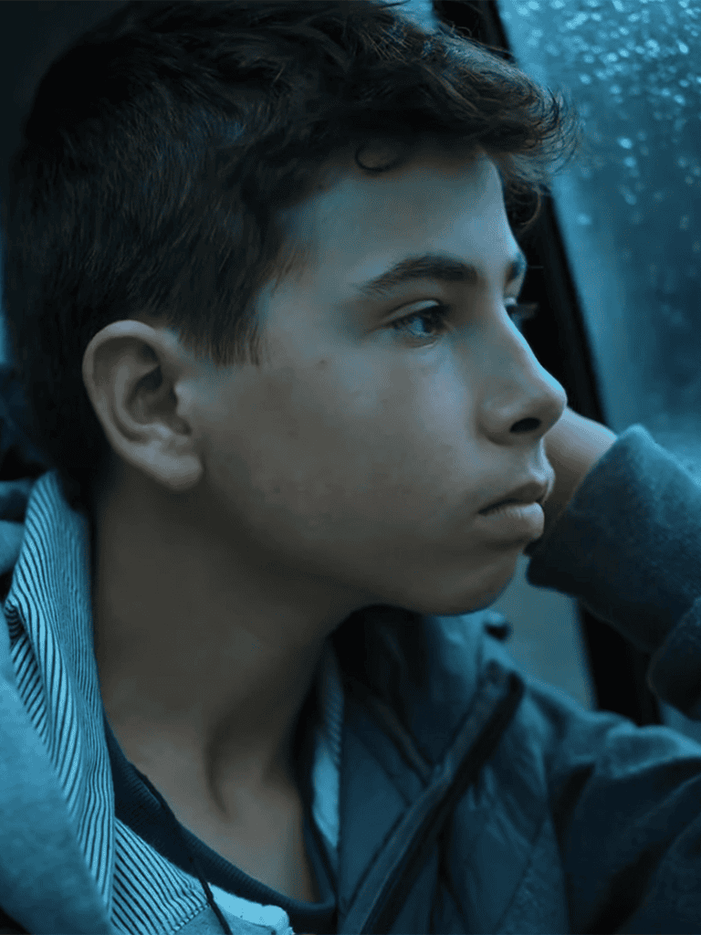 Young man sitting in car, looking thoughtfully outside, casual clothing, moody atmosphere.