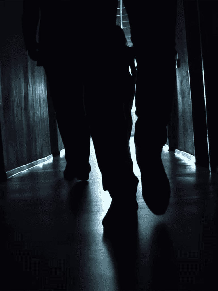 Silhouette of a person walking through a dimly lit corridor with dramatic shadows and lighting.